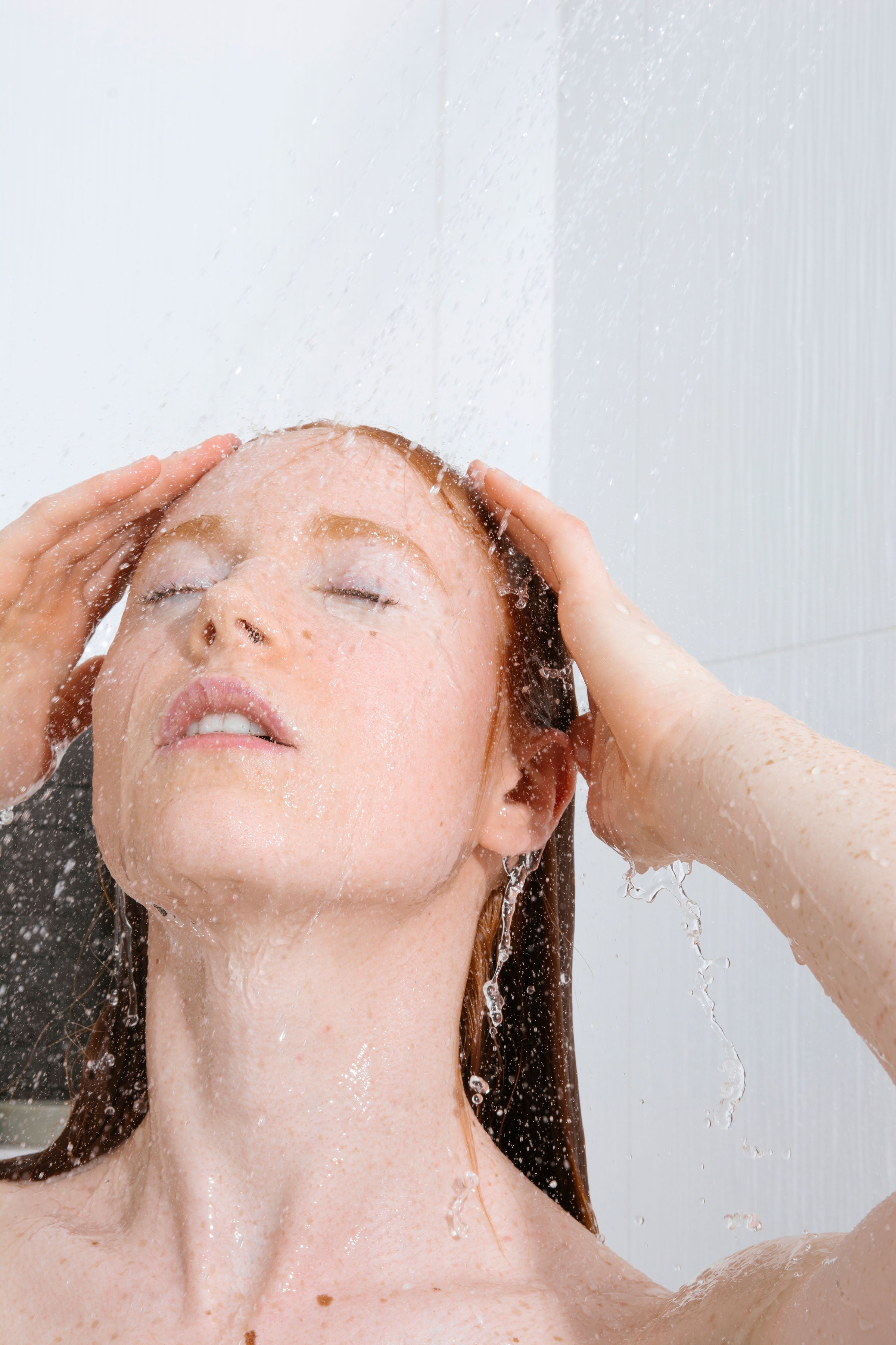 A woman with wet, reddish-brown hair washing her face under running water, with her hands in her hair, set against a light gray tiled background. Rising the hair mask out.